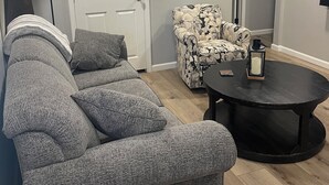 Una sala de estar con un sofá modular gris, un sillón estampado, una mesa de centro redonda y un ventilador de techo.