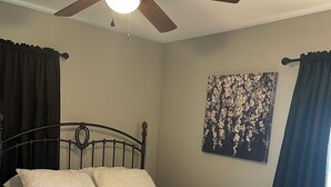 Un dormitorio con cama, ventilador de techo, un cuadro y cortinas.