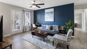 Un salon avec un mur d’accent bleu foncé, un canapé gris, une table basse en bois, un fauteuil à motifs et un ventilateur de plafond.