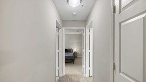 Un couloir étroit aux murs blancs et au plancher en bois mène à une chambre à coucher équipée d’un lit et d’une table de chevet.