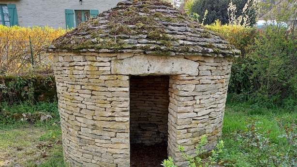 A stone structure with a small opening, surrounded by greenery and a building in the background. - Maison Vacances Pour 4 Personnes (Mont-Saint-Jean)