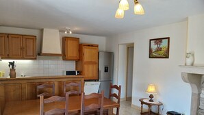 A kitchen with wooden cabinets, a dining table with chairs, a refrigerator, and a small table with a lamp. - Maison Vacances Pour 4 Personnes (Mont-Saint-Jean)