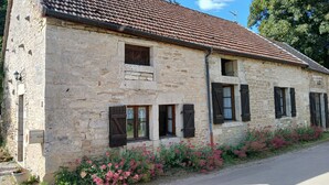 A stone building with a tiled roof, multiple windows with shutters, and a flower bed along the side. - Maison Vacances Pour 4 Personnes (Mont-Saint-Jean)