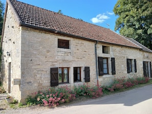A stone building with a tiled roof, multiple windows with shutters, and a flower bed along the side. - Maison Vacances Pour 4 Personnes (Mont-Saint-Jean)