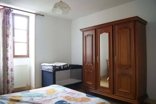 Une chambre avec une armoire en bois, un berceau, une fenêtre avec des rideaux et un luminaire au plafond.