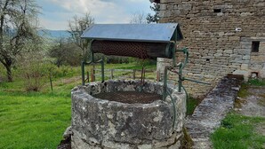 A stone well with a metal structure on top, surrounded by grass and a stone building in the background. - Maison Vacances Pour 4 Personnes (Mont-Saint-Jean)
