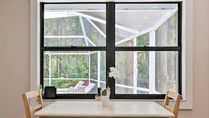 Ein Zimmer mit großem Fenster, einem weißen Tisch, zwei Holzstühlen, einer kleinen Topfpflanze und einer Uhr auf der Fensterbank.