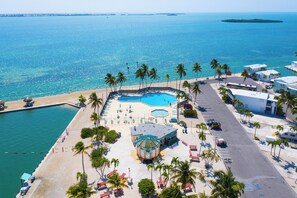 An aerial view of a resort with a large swimming pool, beach area, and palm trees.