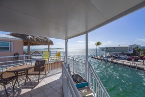 Un balcon avec une table et des chaises, un toit de chaume et une vue sur la mer et les bâtiments.