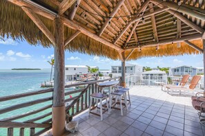 A covered outdoor area with a wooden deck, chairs, and a view of the sea and buildings.