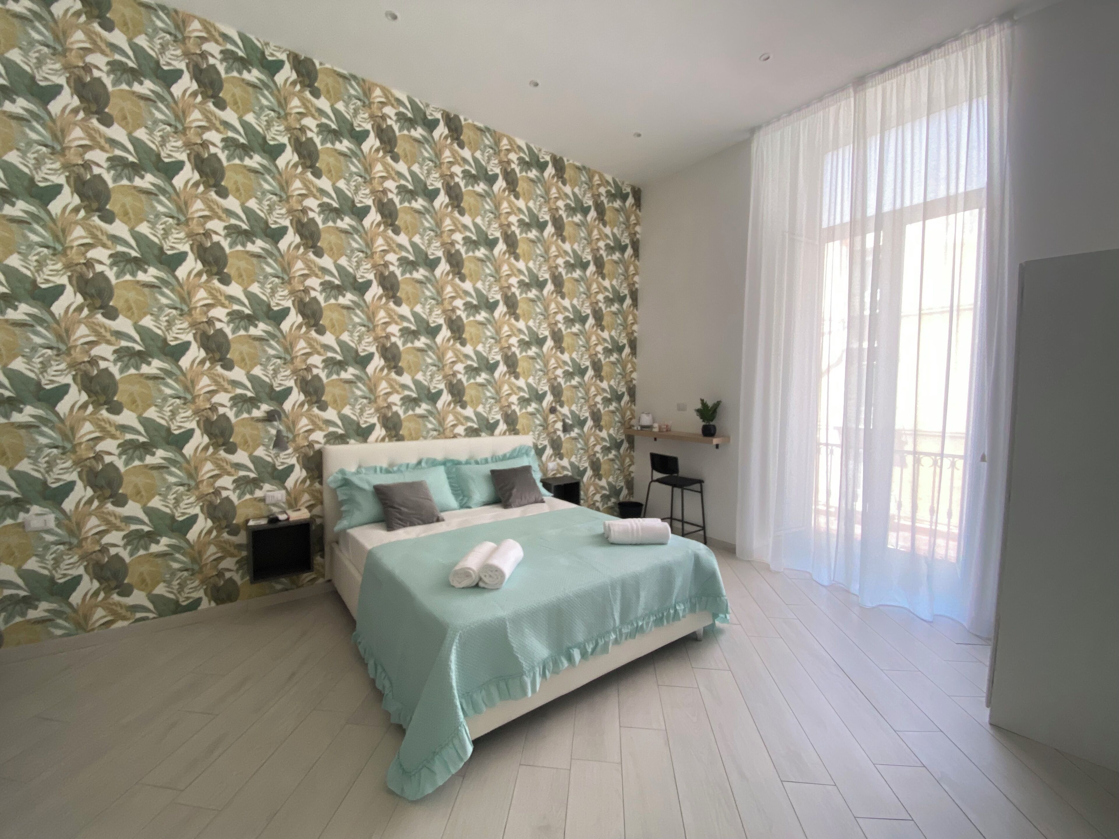 Ein Schlafzimmer mit floraler Tapete, einem Bett mit türkisner Bettdecke und einem großen Fenster mit transparenten Vorhängen.