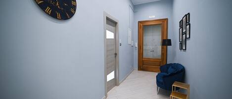 Een hal met een wandklok, een deur, een blauwe stoel en een klein houten plankje.