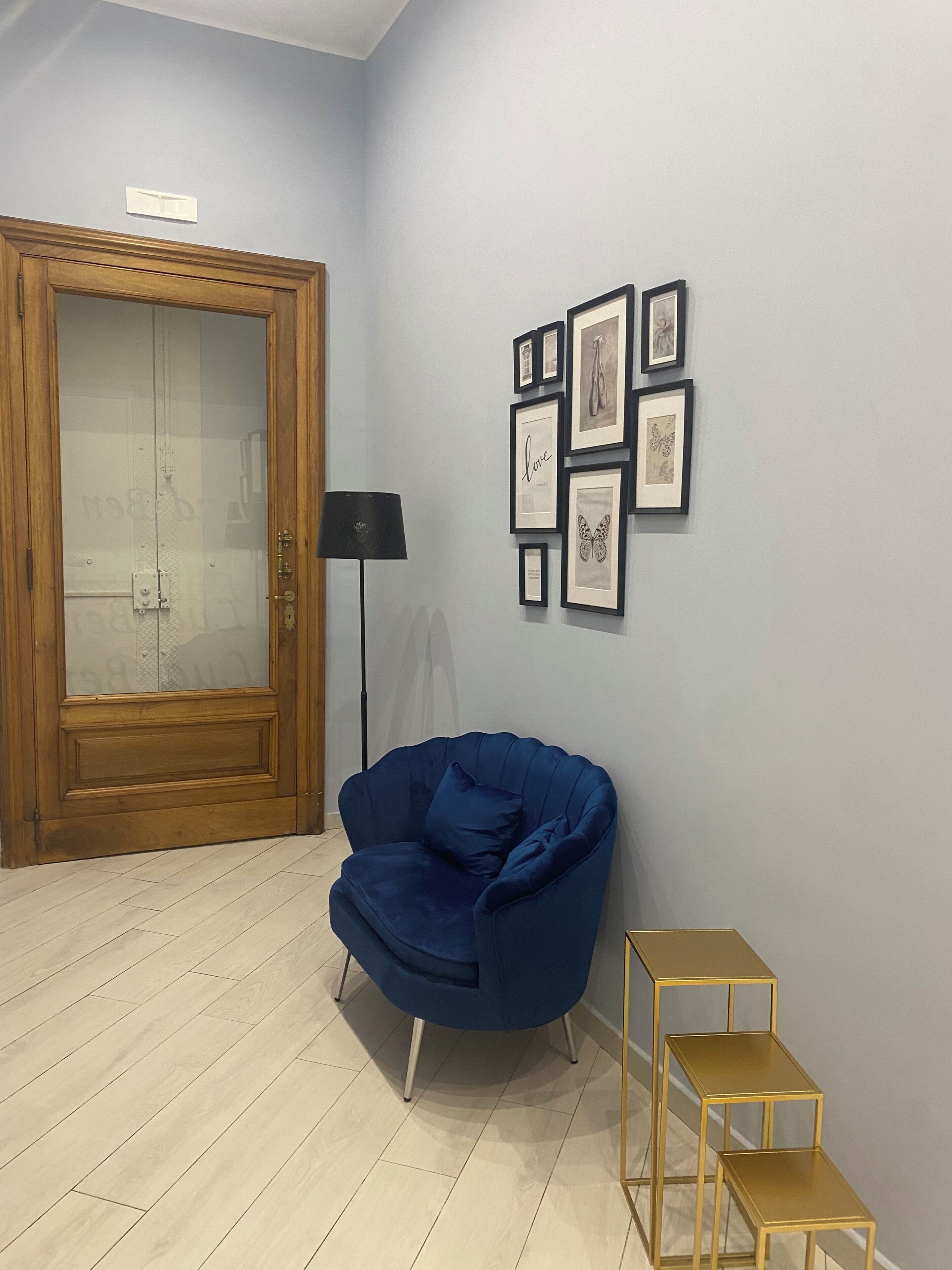 A room with a wooden door, a blue armchair, a floor lamp, and a wall with framed pictures.