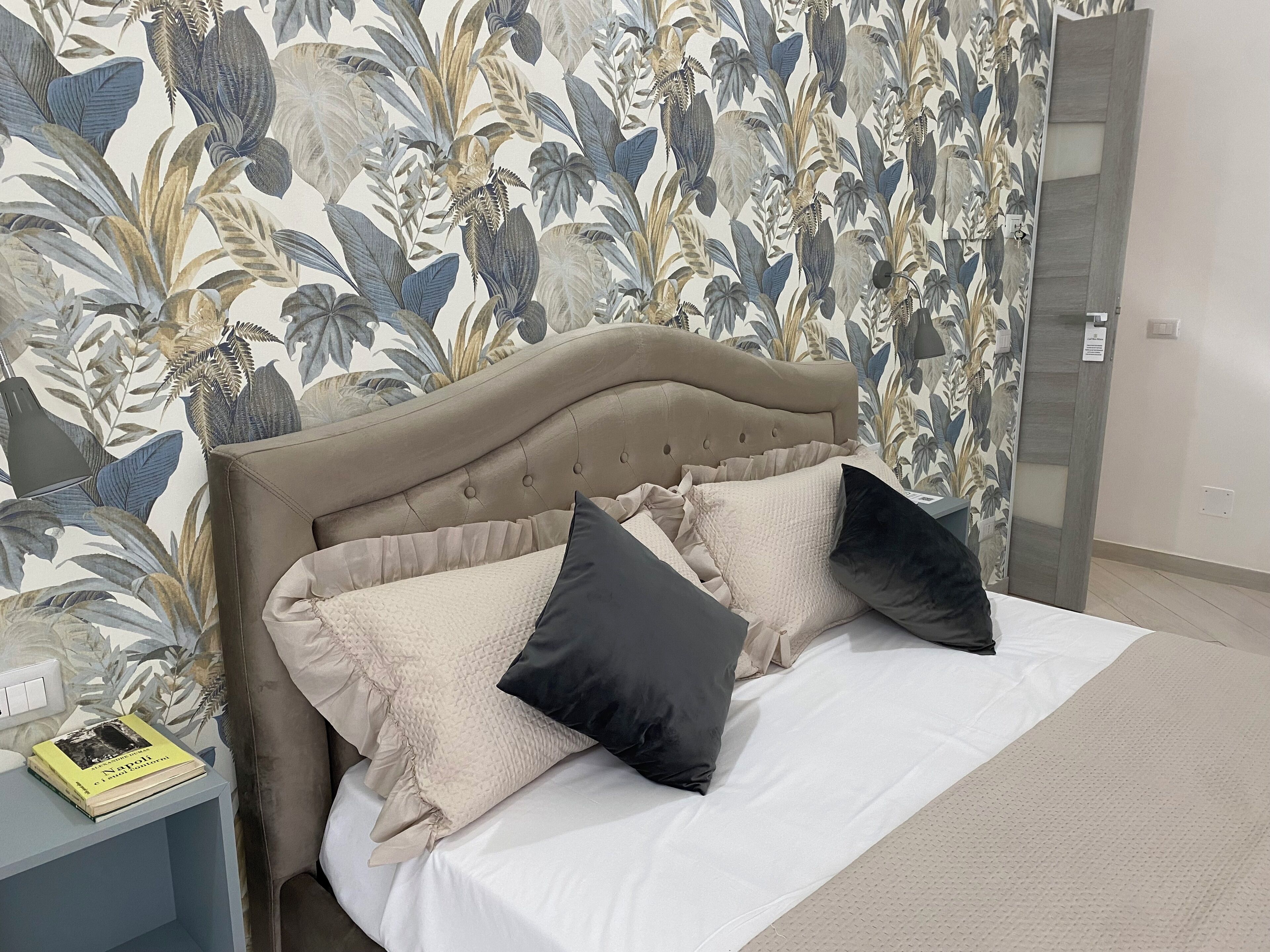Een slaapkamer met een bed, twee kussens, een nachtkastje en bloemenbehang.