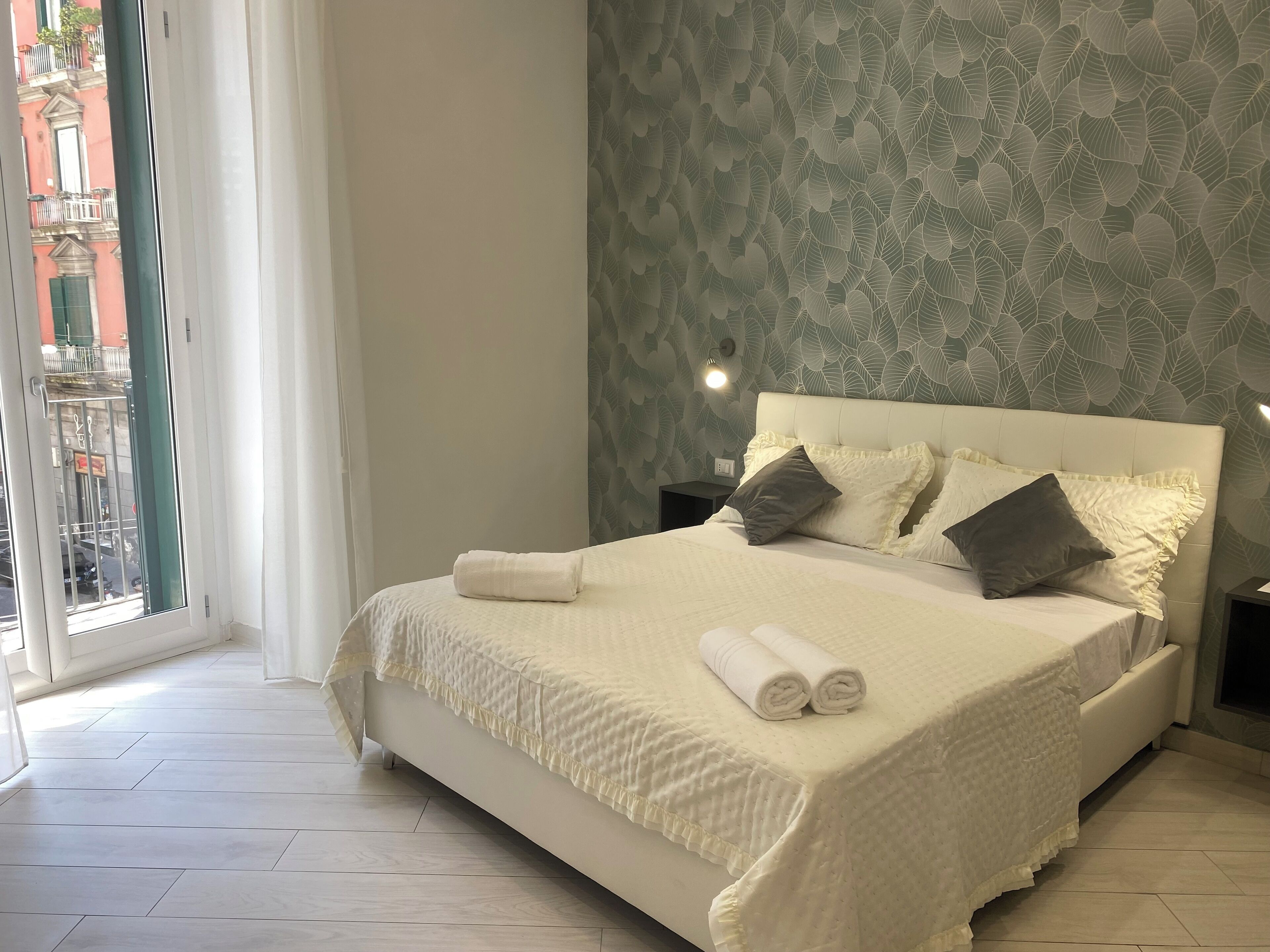 A neatly made bed with pillows and a rolled towel, a bedside table with a lamp, and a patterned wall.