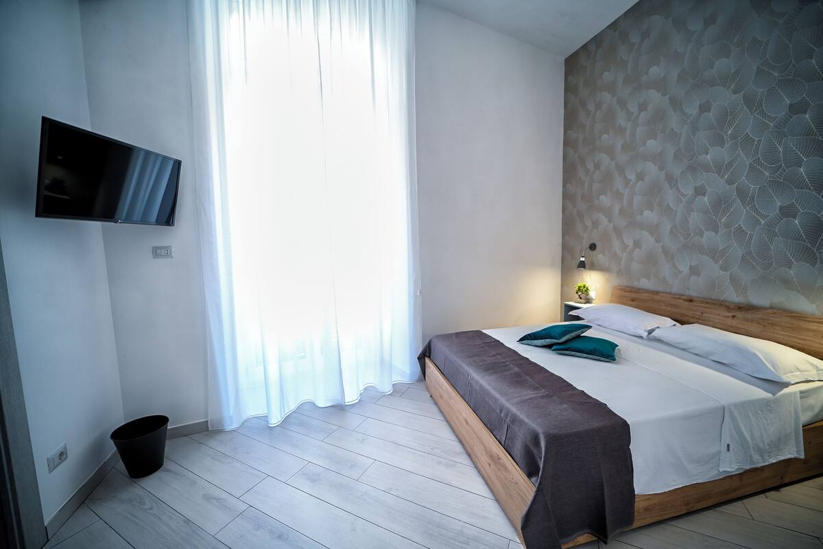 Ein Schlafzimmer mit Bett, Wandfernseher, Fenster mit Vorhängen und einer Wand mit Muster.