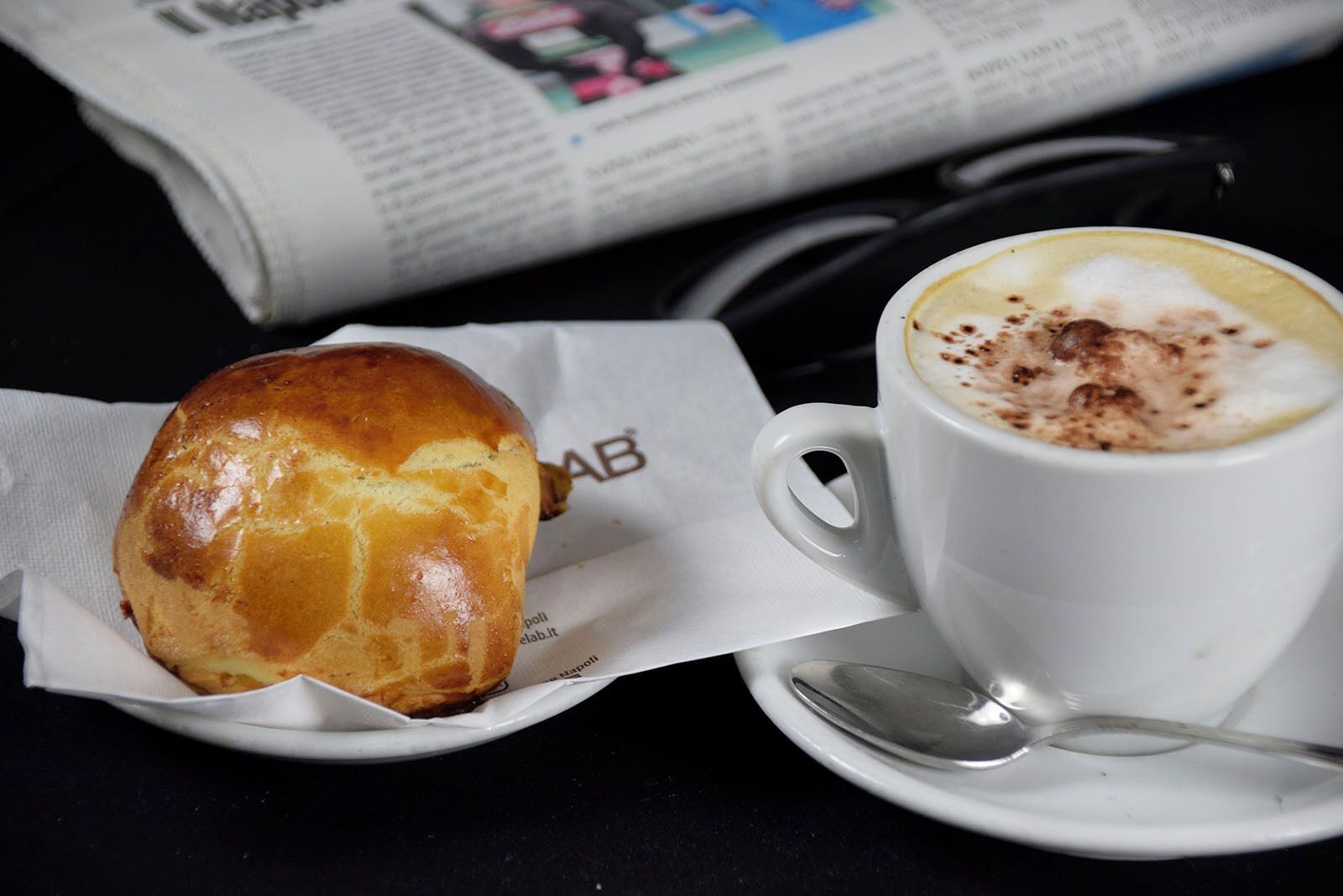A sandwich and a cup of coffee on a table with a newspaper and glasses.
