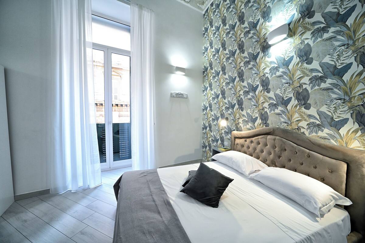 Ein Schlafzimmer mit einem großen Bett, Blick ins Freie durch eine Schiebetür und floralem Wandpapier.