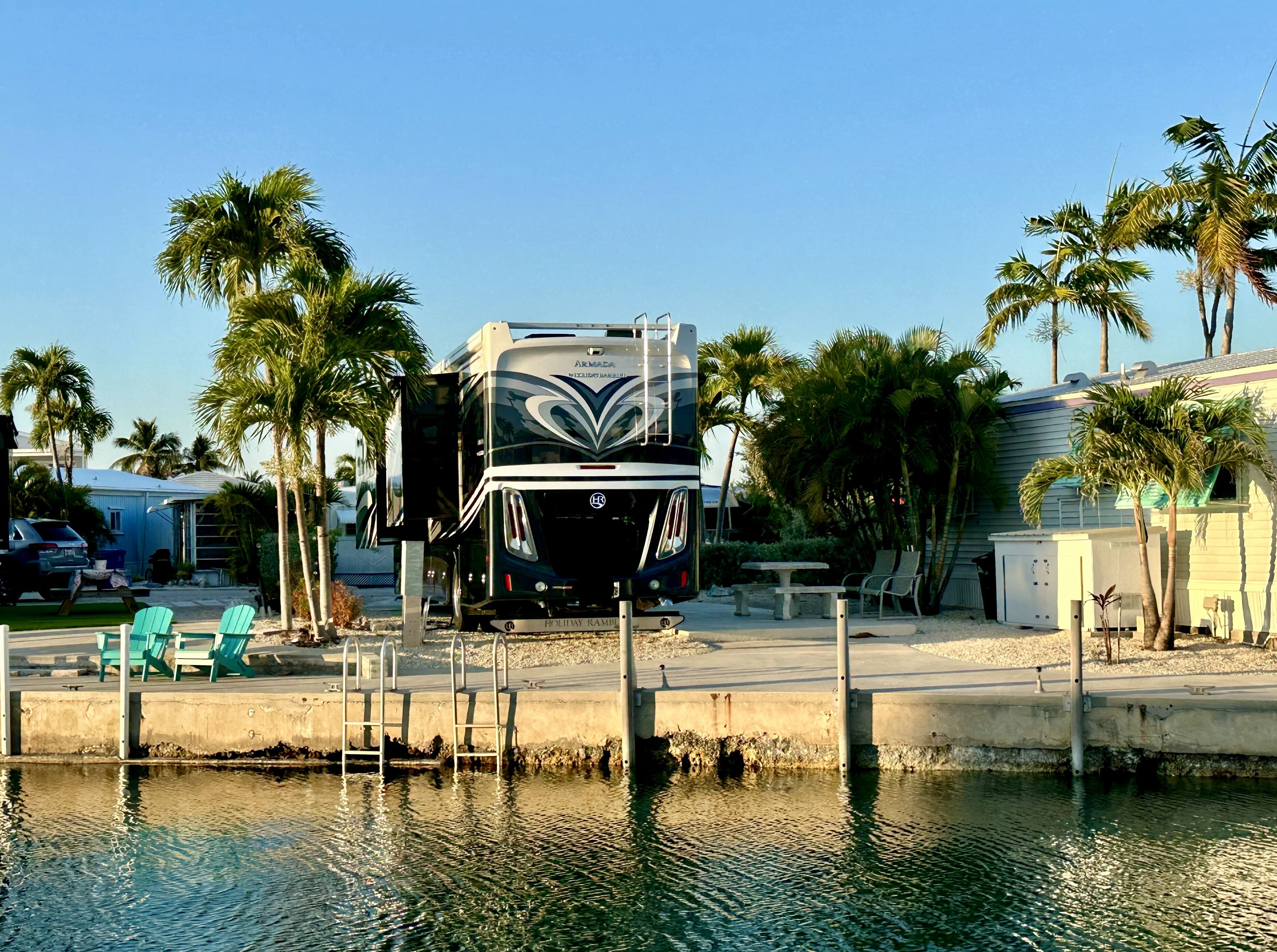 A large RV parked by a waterfront with palm trees and a small dock.