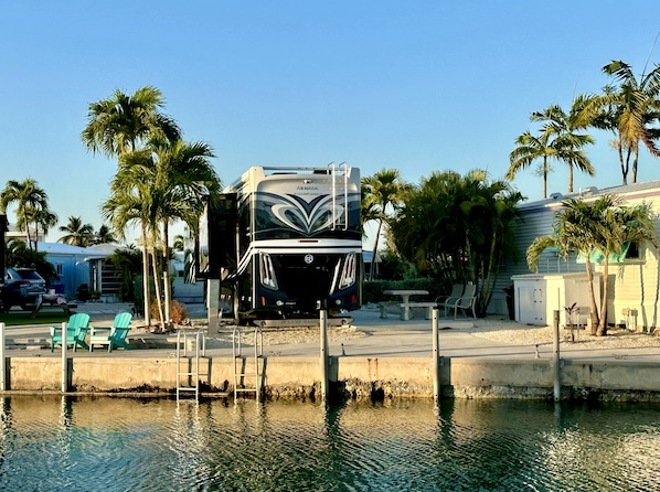 Un grand camping-car est garé au bord de l’eau, à proximité de palmiers et d’un petit quai.