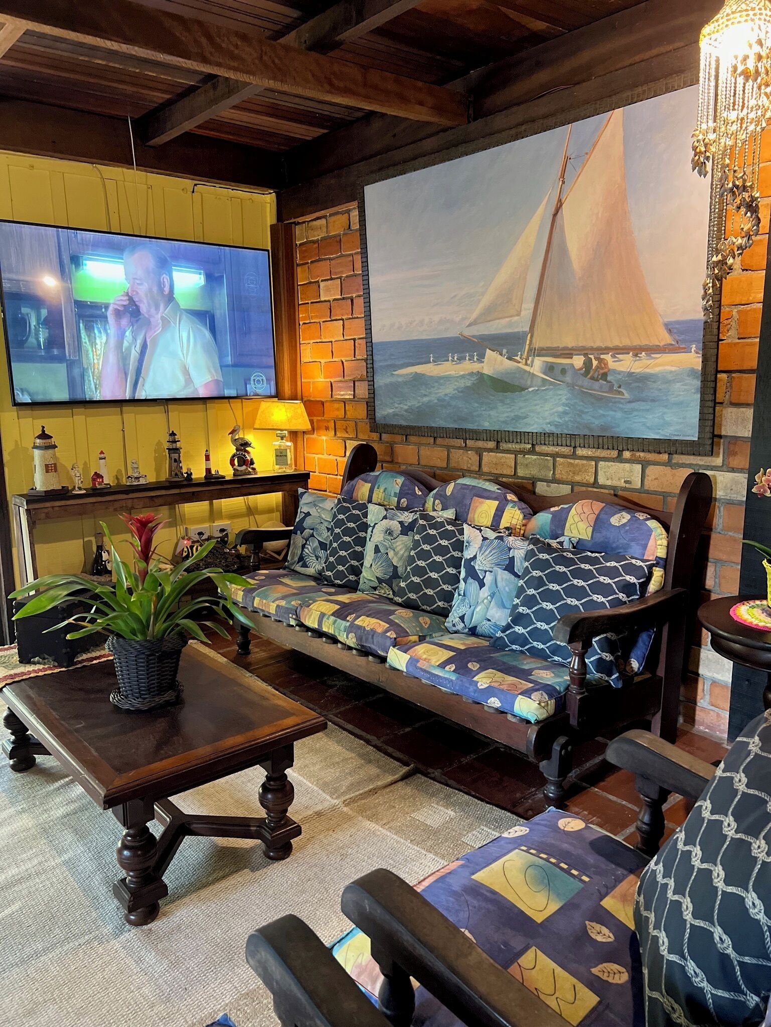 A cozy living room with a wooden ceiling, a television mounted on the wall, a large sailboat painting, a patterned sofa, and a small table with a plant.