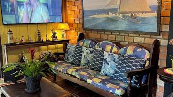 A cozy living room with a wooden ceiling, a television mounted on the wall, a large sailboat painting, a patterned sofa, and a small table with a plant.