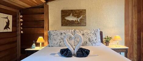 Habitación de cabaña de madera con cama, dos lámparas, decoración de tiburón en la pared y un arreglo de toallas con forma de corazón.