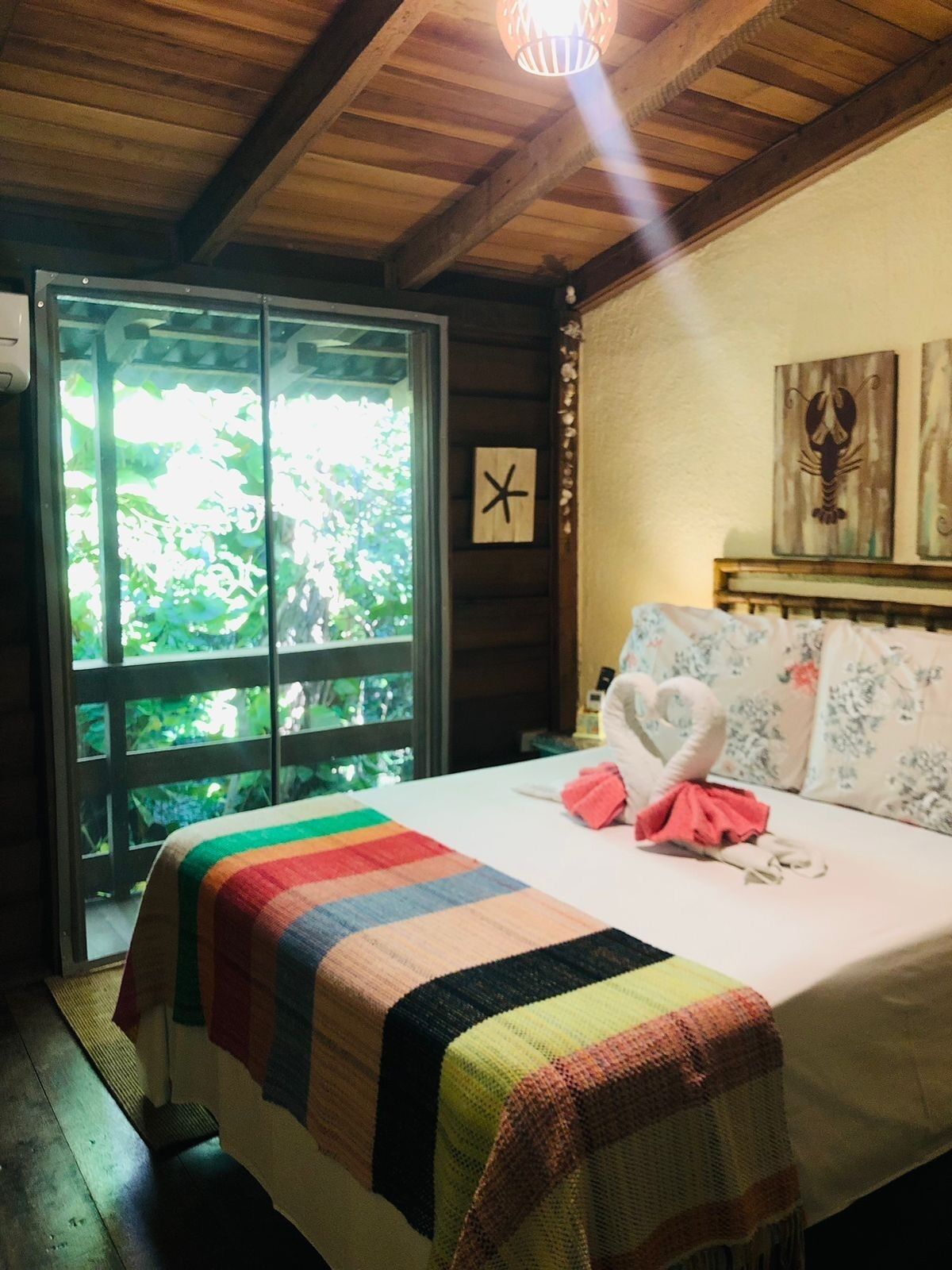 A room with a bed, a window with a view of greenery, a wooden ceiling, and a wall clock.