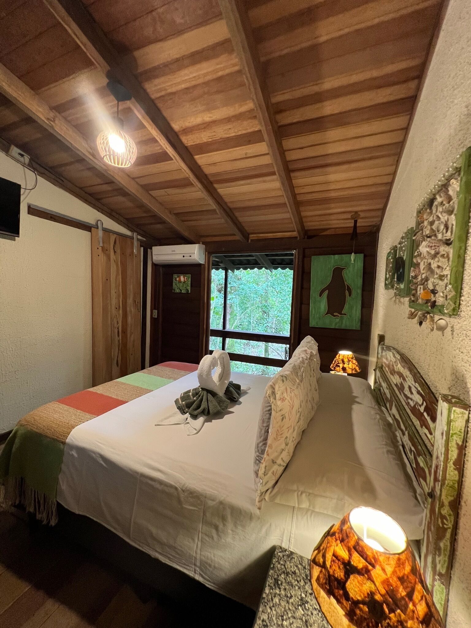 Una habitació acollidora amb sostre de fusta, un llit amb edredó blanc, una decoració mural verda i una finestra amb vista a vegetació.
