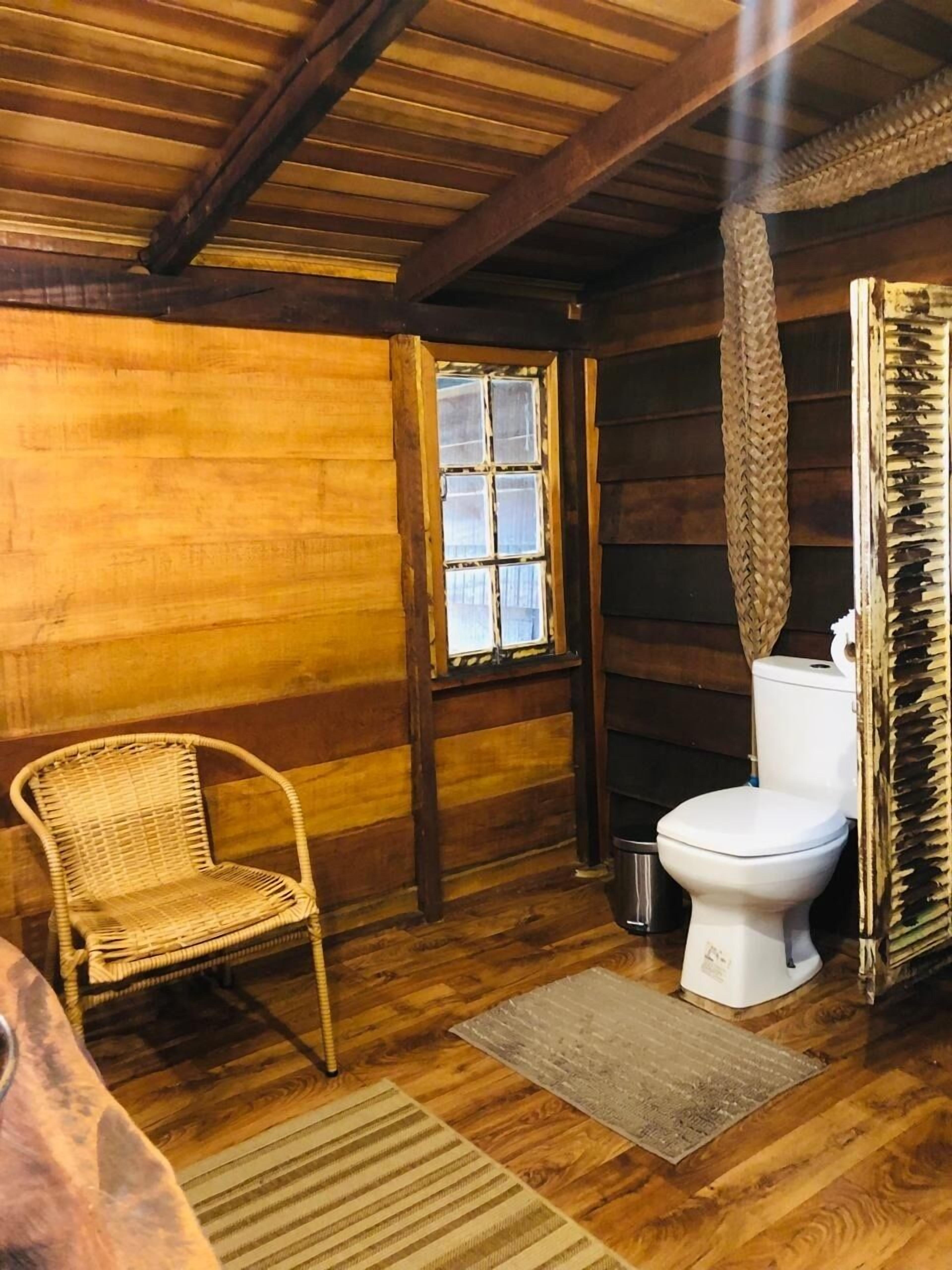 A rustic wooden cabin interior with a single wicker chair, a white toilet, and a window with a woven basket hanging above it.