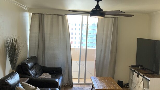 Una sala moderna con un sofá de cuero, una mesa de centro de madera, un televisor y un balcón con cortinas.