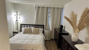 Un dormitorio con cama, mesitas de noche, cómoda, radiador y ventana con cortinas.
