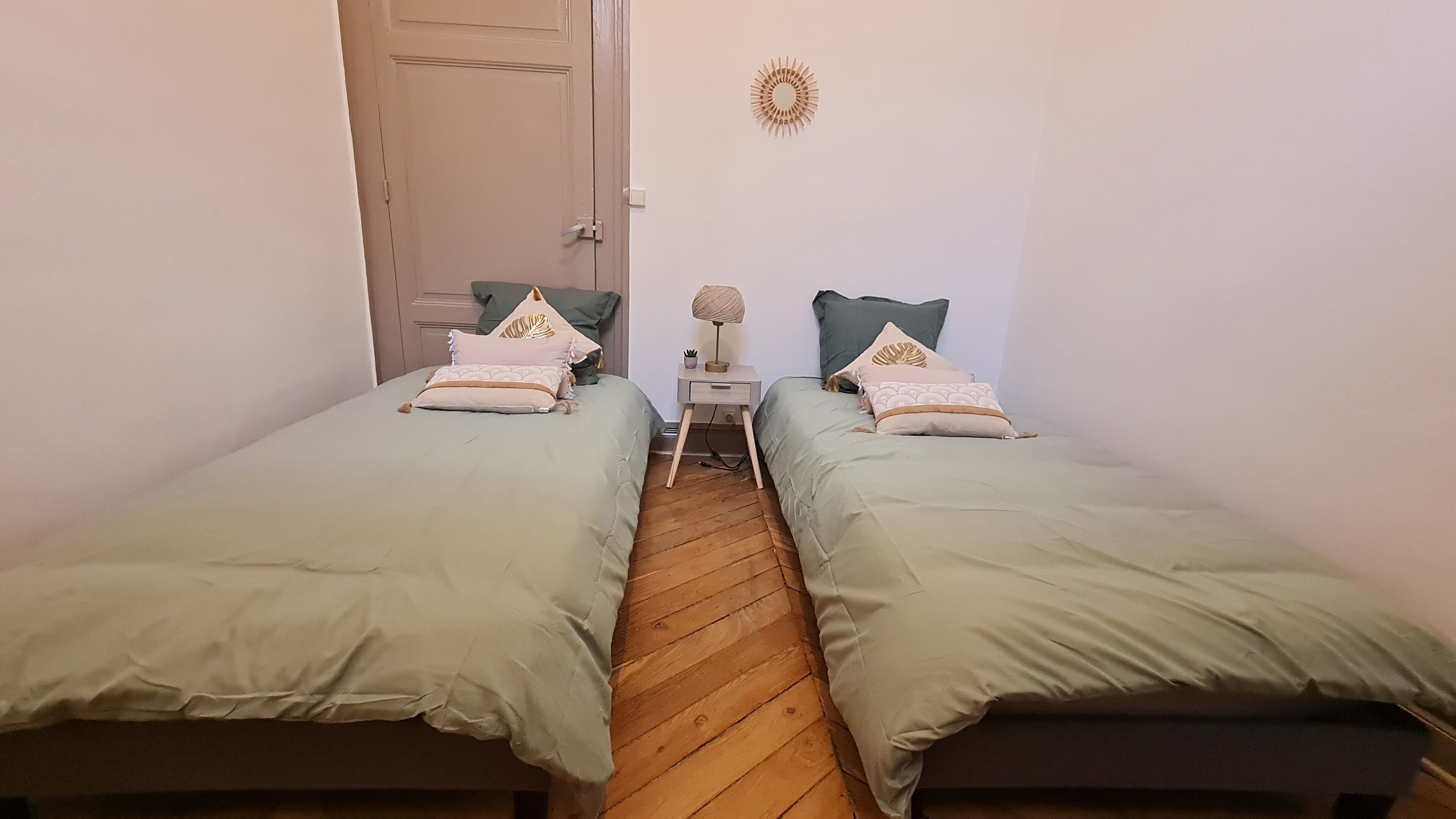 Et lille, smalt værelse med to enkeltsenge, trægulv, et lille bord med en lampe og en dør.