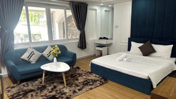 Ett modernt hotellrum med en blå soffa, en vit säng med ett vadderat täcke, ett litet runt bord med en vas med blommor och ett trägolv.
