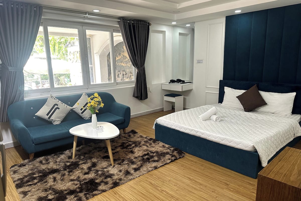 A modern hotel room with a blue sofa, a white bed with a quilted duvet, a small round table with a vase of flowers, and a wooden floor.