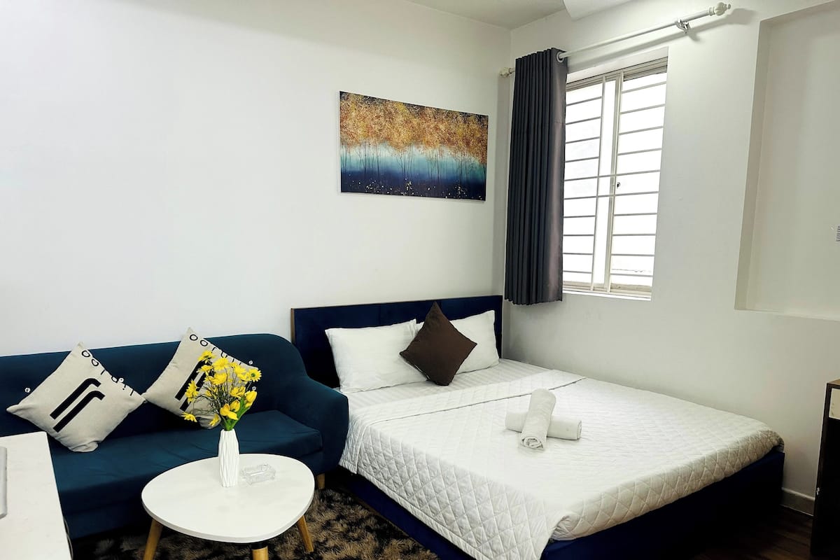 A hotel room with a bed, a blue sofa, a small white table with a vase of flowers, and a painting on the wall.