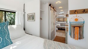 A bedroom with a bed, a window with curtains, a wall-mounted dreamcatcher, and a door leading to another room.