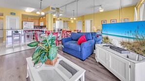A living room with a blue sofa, a coffee table with plants, a TV, and a view of the beach through a large window. - Sunrise Beach 2107 (Panama City Beach)
