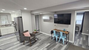 Una cocina moderna con armarios blancos, un refrigerador de acero inoxidable y un horno empotrado. Hay un comedor con una mesa de madera y dos sillas azules. Un televisor de pantalla plana está montado en la pared sobre la mesa de comedor.