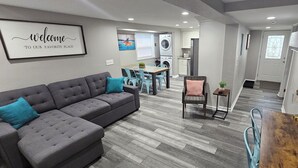 Una sala de estar moderna con un sofá seccional gris, un comedor con mesa de madera y una cocina al fondo.