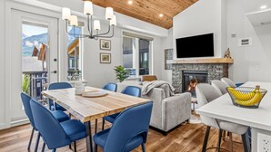 A modern living room with a fireplace, a dining area with blue chairs, and a TV mounted on the wall. - Three Sisters Skyline | Designer Penthouse (Canmore)