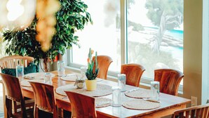 Zona pranzo con tavolo in legno apparecchiato per sei persone, circondato da sedie in legno. Sul tavolo c'è una pianta in vaso e la stanza ha ampie finestre.