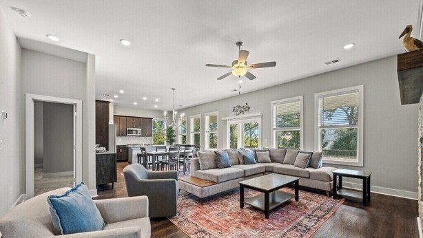 A spacious living room with a large sectional sofa, a coffee table, and a patterned rug. There is a dining area with a table and chairs, and a kitchen in the background.