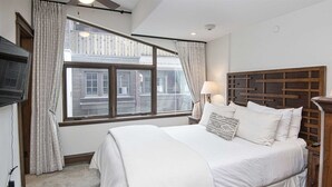 A bedroom with a large bed, a wooden headboard, a window with curtains, a nightstand with a lamp, and a TV mounted on the wall. - 5 Bedroom in the Heart of Vail Village (Vail)