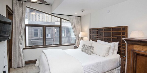 A bedroom with a large bed, a wooden headboard, a window with curtains, a nightstand with a lamp, and a TV mounted on the wall. - 5 Bedroom in the Heart of Vail Village (Vail)