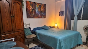 Un dormitorio con cama, una silla, una mesa, un armario, un cuadro, una lámpara y un ventilador.