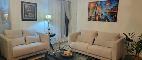 Una sala de estar con dos sofás color beige, una mesa de centro de vidrio, una araña, un cuadro y una planta en maceta.