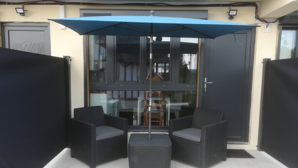 A patio area with two wicker chairs and a table, a blue umbrella, and a glass door leading to an interior space. - Location de Courte Durée Studio Meublé (Le Trait)