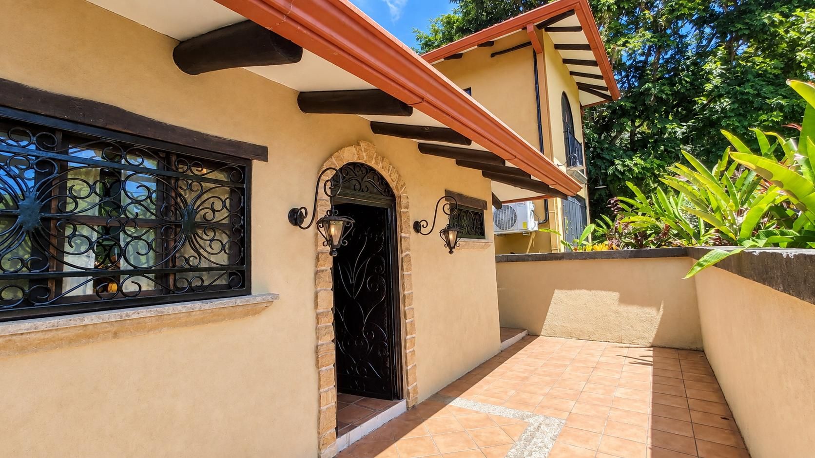 A building with a tiled patio, a black door with a stone arch, and a window with decorative iron bars.