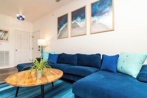 A living room with a blue sofa, a wooden coffee table with a plant, and three framed artworks on the wall. - Spacious, modern 2-bed/2-bath condo in marvelous Rehoboth Beach! (Rehoboth Beach)
